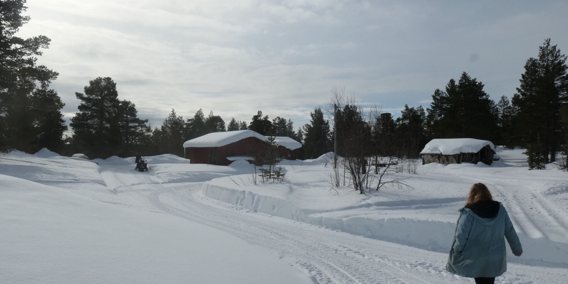 Person in a blue coat walks on a snow-covered path towards rustic buildings surrounded by snowy trees under a cloudy sky.