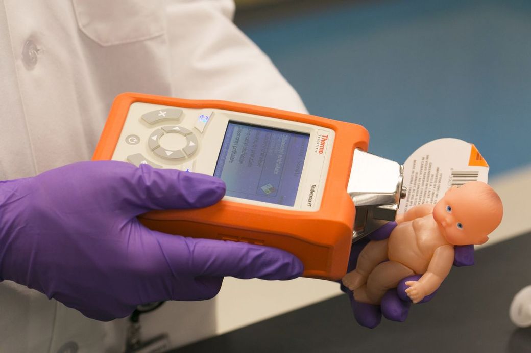 Een man in laboratoriumjas en met plastic handschoenen houdt een meetapparaat bij een in beslag genomen speelgoedpoppetje.