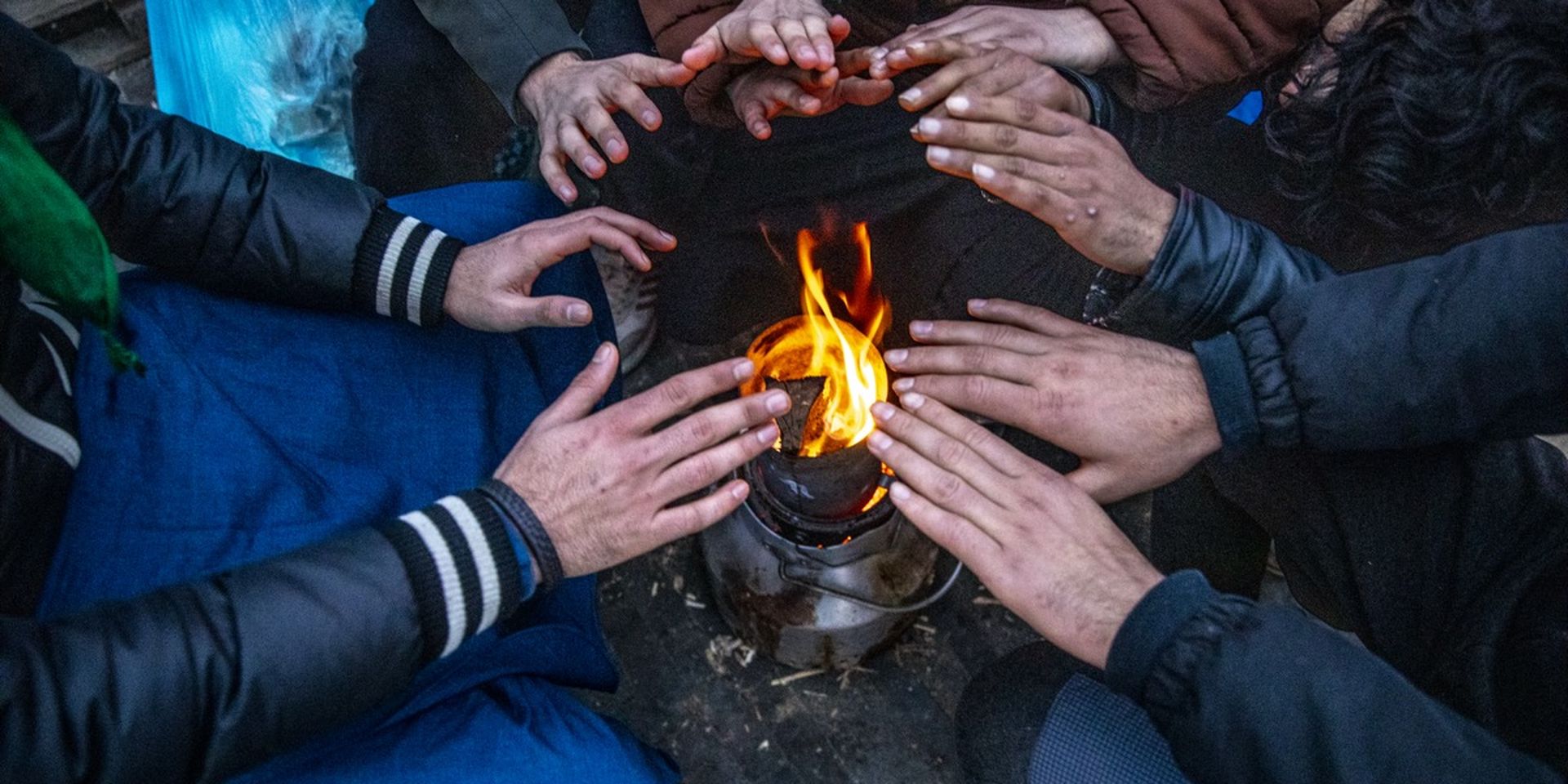 People gather around a small open fire outdoors, warming their hands in the cold.