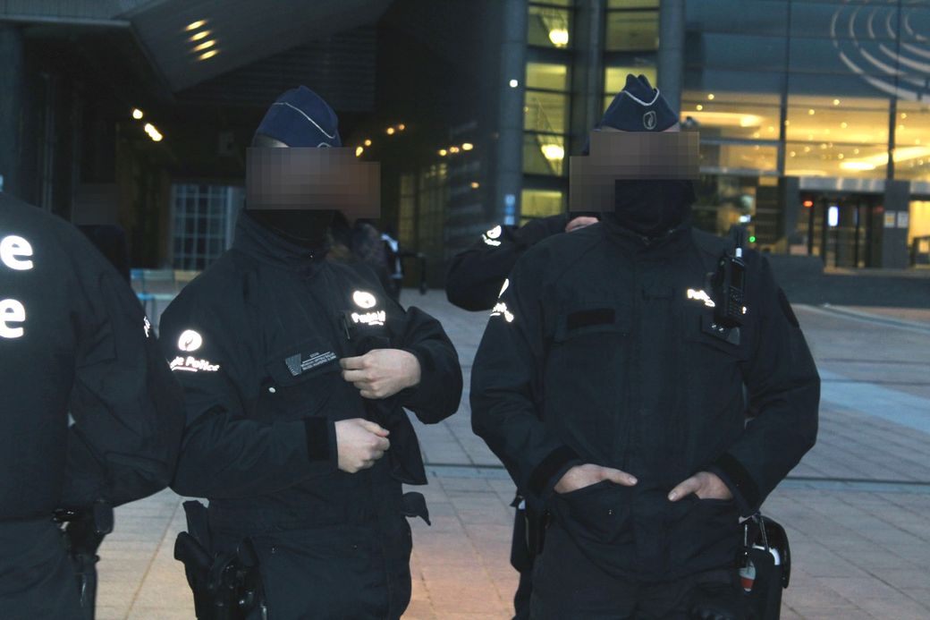 Two police officers in uniform and caps stand outside a building, partially covering their faces with black masks.