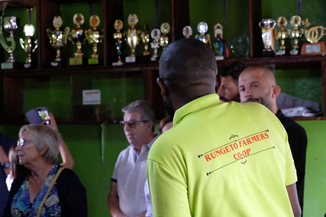 A group of people stand in front of shelves filled with trophies. A man faces them wearing a bright shirt labeled "Rungeto Farmers Co-op."