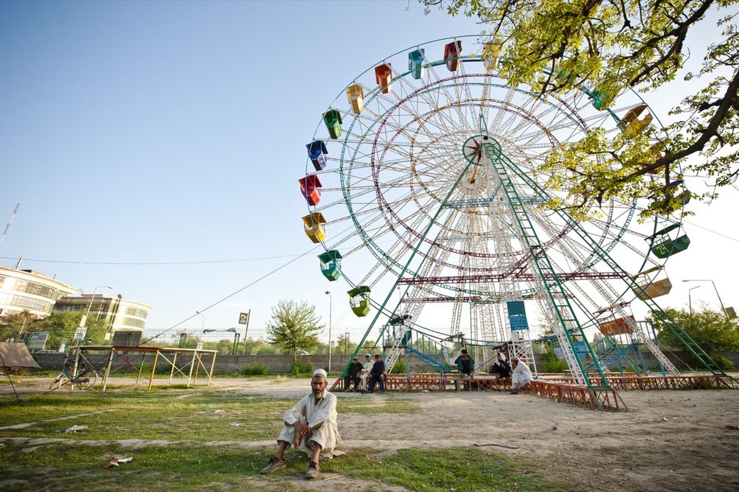 Man sitting near a large Ferris wheel with colorful carriages, surrounded by trees and buildings under a clear blue sky.