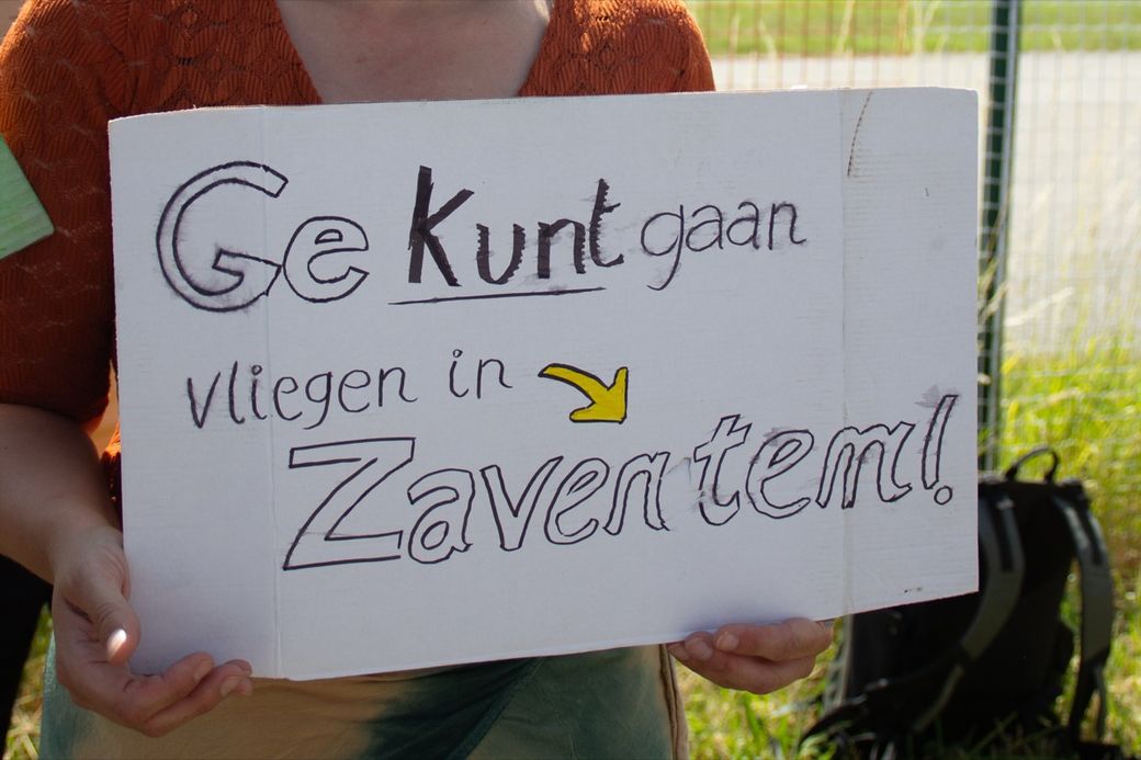 Person holding a handwritten sign in Dutch, "Ge kunt gaan vliegen in Zaventem!" with an arrow pointing to "Zaventem," outside.