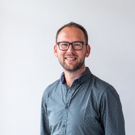 Smiling man with glasses in a blue polka dot shirt against a plain white background.