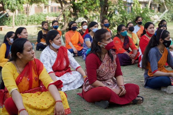 A group of women sit on the grass outdoors, wearing colorful traditional attire and face masks. Trees and a building are visible in the background.