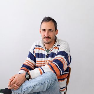 Man in a striped sweater and jeans sits on a chair, crossing his legs, against a plain white background.