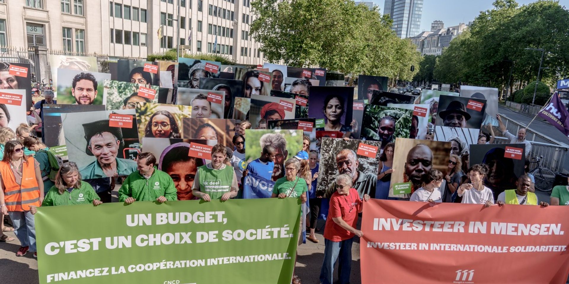People holding large portraits and banners advocating for international cooperation in a demonstration outside modern buildings.
