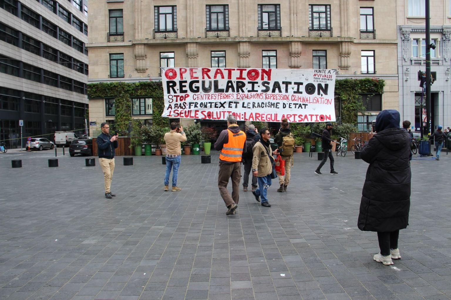 People in a plaza holding a large banner reading "Operation Regularisation" with messages against deportation and state racism.