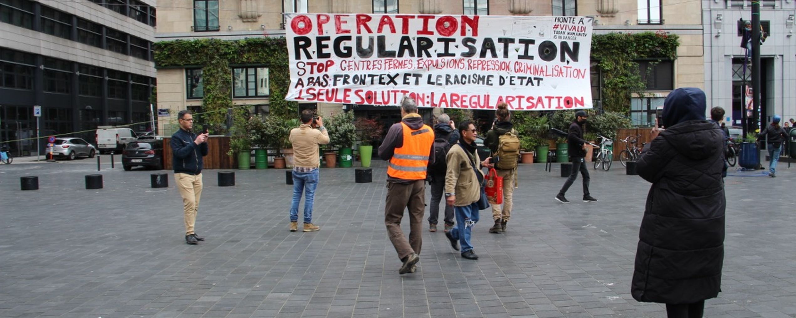 People in a plaza holding a large banner reading "Operation Regularisation" with messages against deportation and state racism.