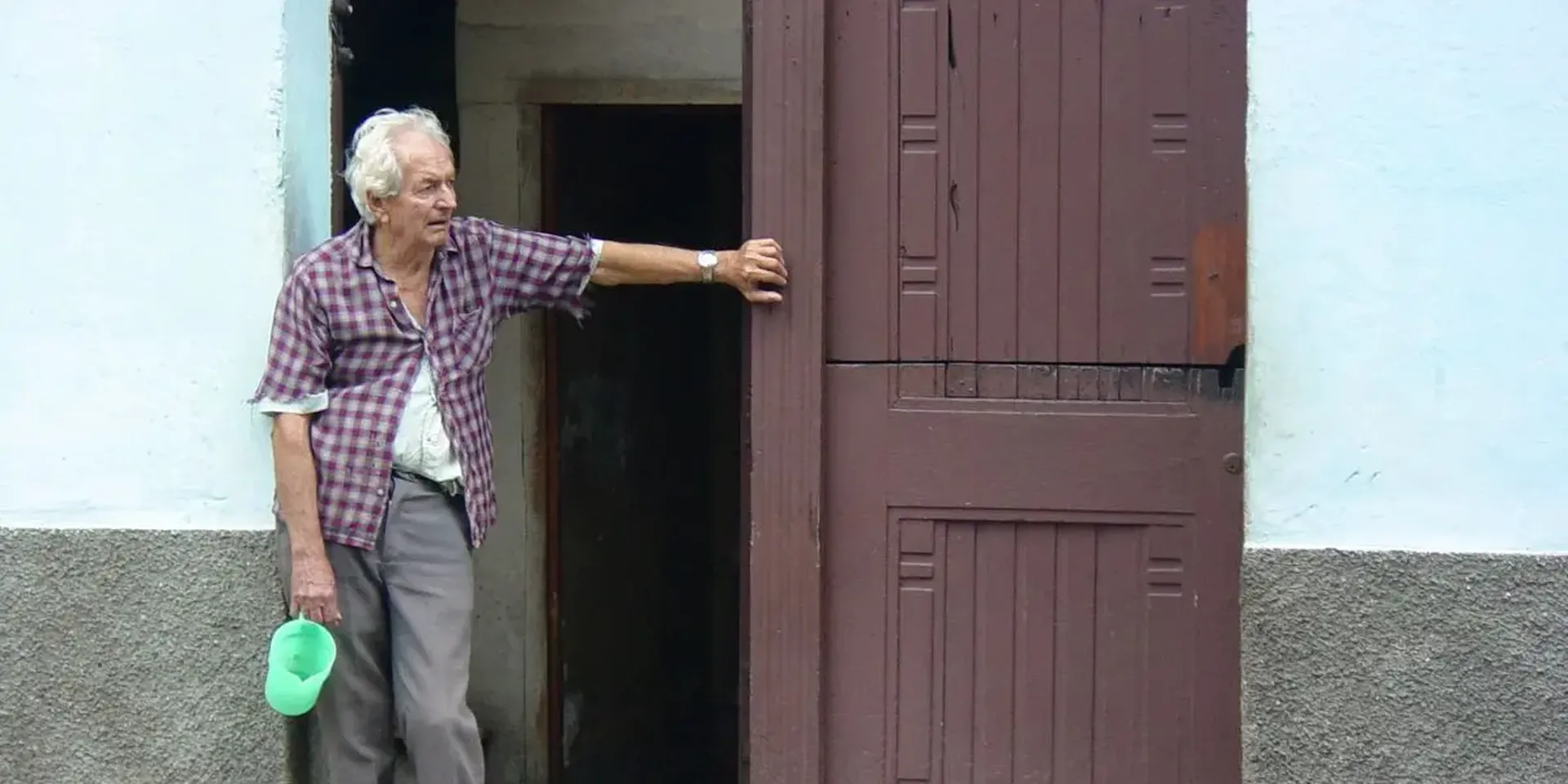Een oudere man leunt tegen de portiek van zijn woning met een lege waterkan in de hand