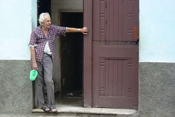Een oudere man leunt tegen de portiek van zijn woning met een lege waterkan in de hand