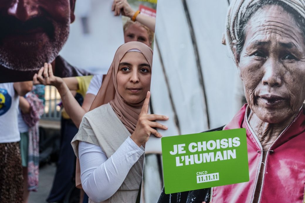 A woman in a pink hijab holds a sign with another image and the text "Je Choisis L'Humain CNCD 11.11.11," standing among others with posters.