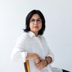 A woman with dark hair and glasses sits on a chair, wearing a white shirt, against a plain white background.