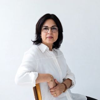 A woman with dark hair and glasses sits on a chair, wearing a white shirt, against a plain white background.