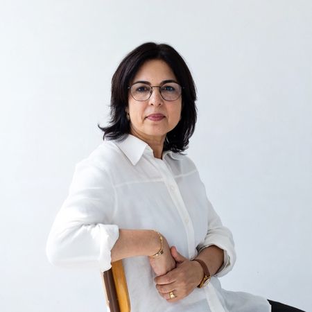 A woman with dark hair and glasses sits on a chair, wearing a white shirt, against a plain white background.