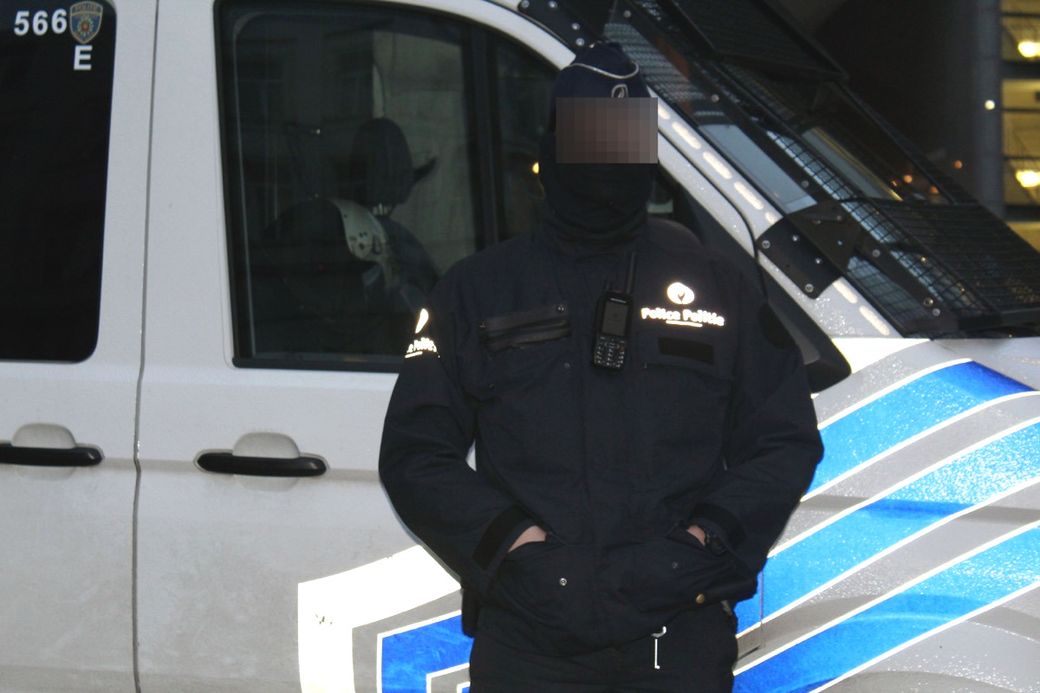 Police officer in uniform standing beside a patrol van, wearing a cap and face covering.
