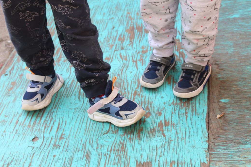 Twee kinderen staan op een houten ondergrond met afgebladderde verf, enkel hun voeten en benen worden getoond.