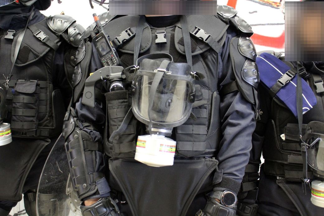 Three people in heavy tactical gear, including gas masks and protective vests, standing close together in a line.