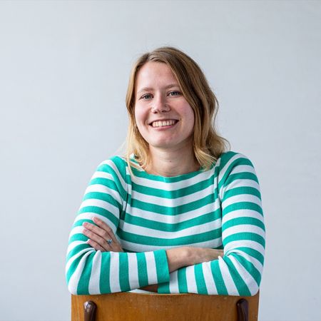 Smiling person in a green and white striped shirt sitting on a chair against a plain background.