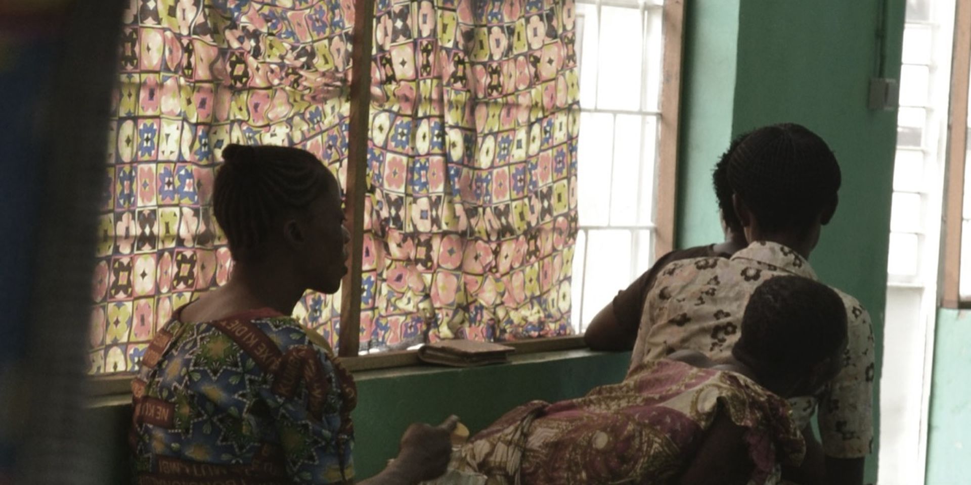 Women in vibrant patterned clothing sit in a room with colorful curtains, one holding a child.