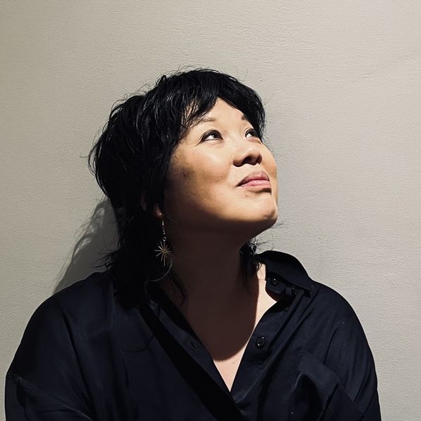 A person with short, dark hair gazes upward thoughtfully, wearing a black shirt and star-shaped earrings against a plain background.