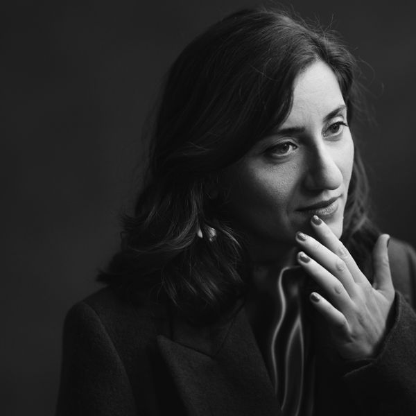 Black and white portrait of a woman with wavy hair, looking thoughtful, hand near her chin, wearing a coat against a dark backdrop.