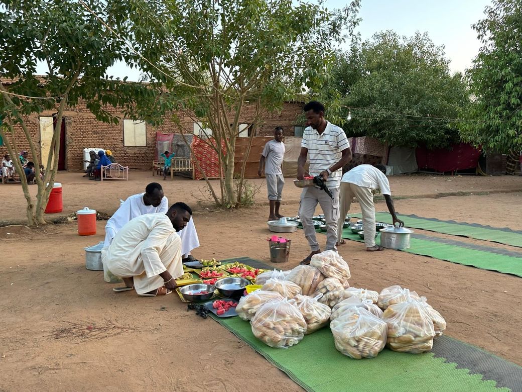People preparing food on mats outdoors, with bags of bread and assorted items, near a brick building and trees.