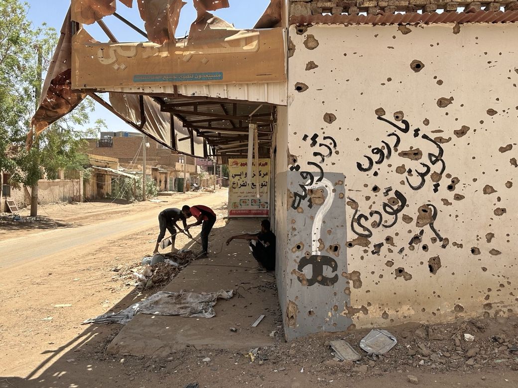 Street scene with two people cleaning near a bullet-riddled wall with graffiti. A damaged sign and debris are visible under a clear sky.