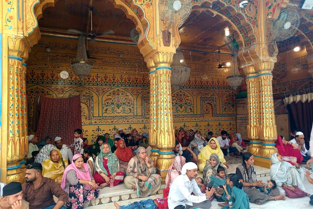 A group of people, including men, women, and children, seated in a vividly decorated, ornate hall with arches and Islamic calligraphy.