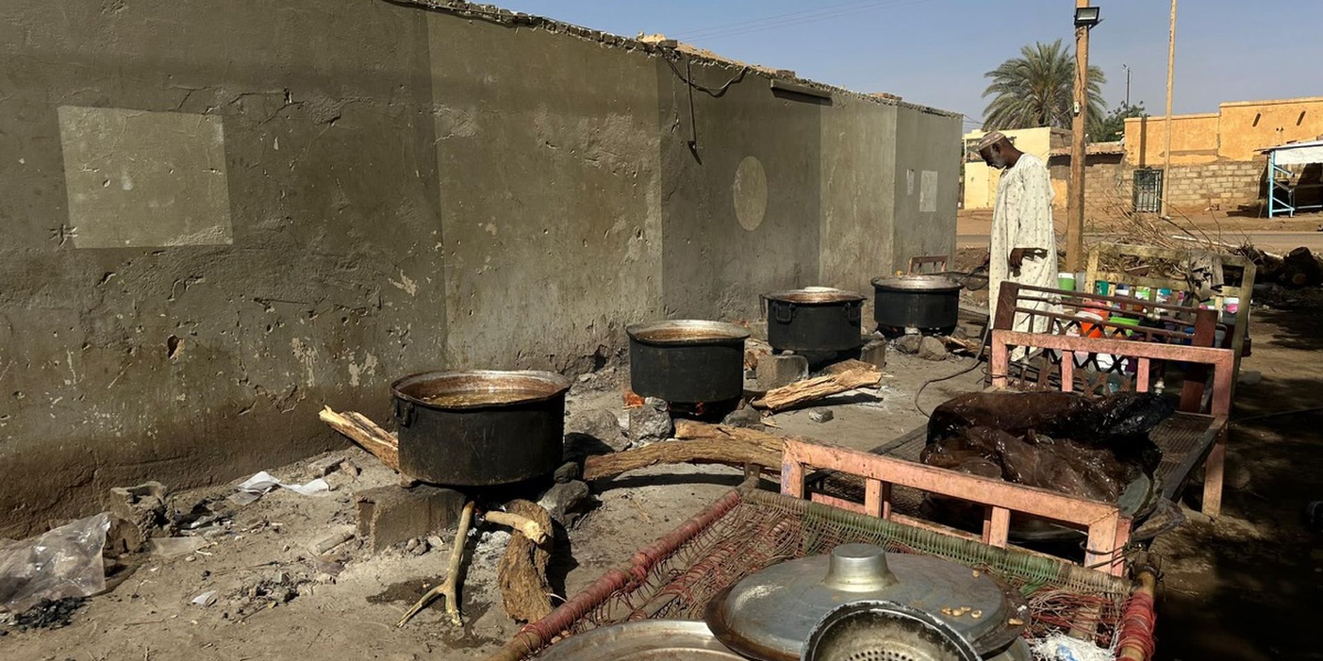 Outdoor cooking setup with large pots on wood fires by a wall. A person stands nearby, with kitchen utensils and a bench in the foreground.