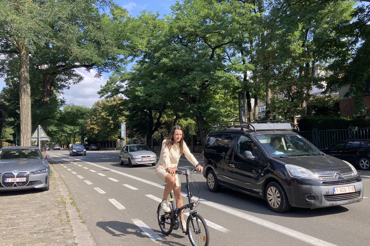 A person in light clothing rides a bicycle on a tree-lined street, with parked cars and moving traffic on a sunny day.