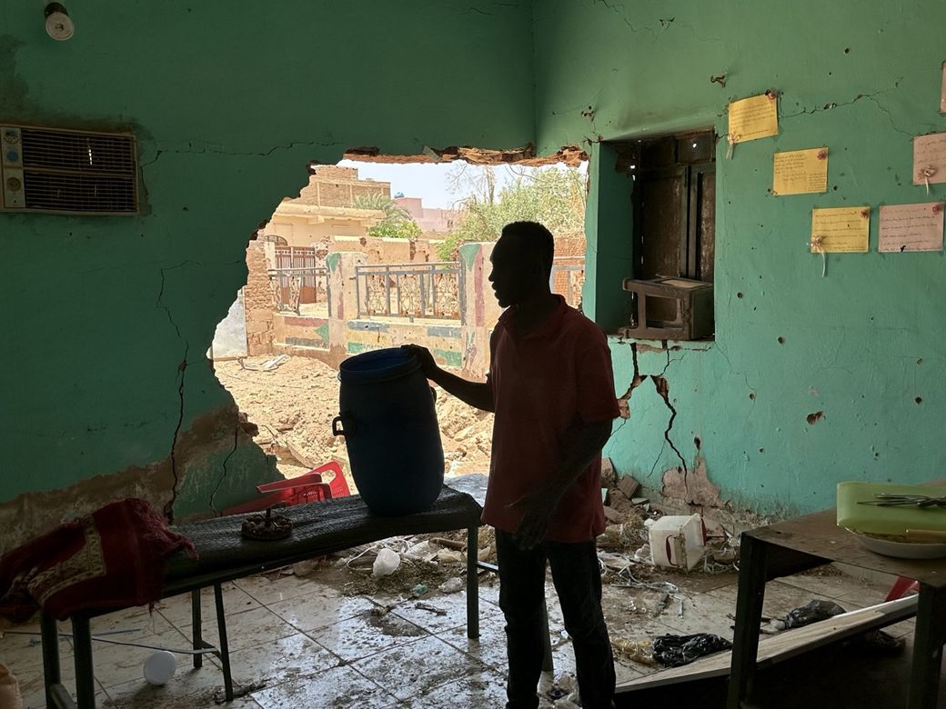 A person stands in a damaged room with a large hole in the wall, holding a blue container. Debris is scattered on the floor.