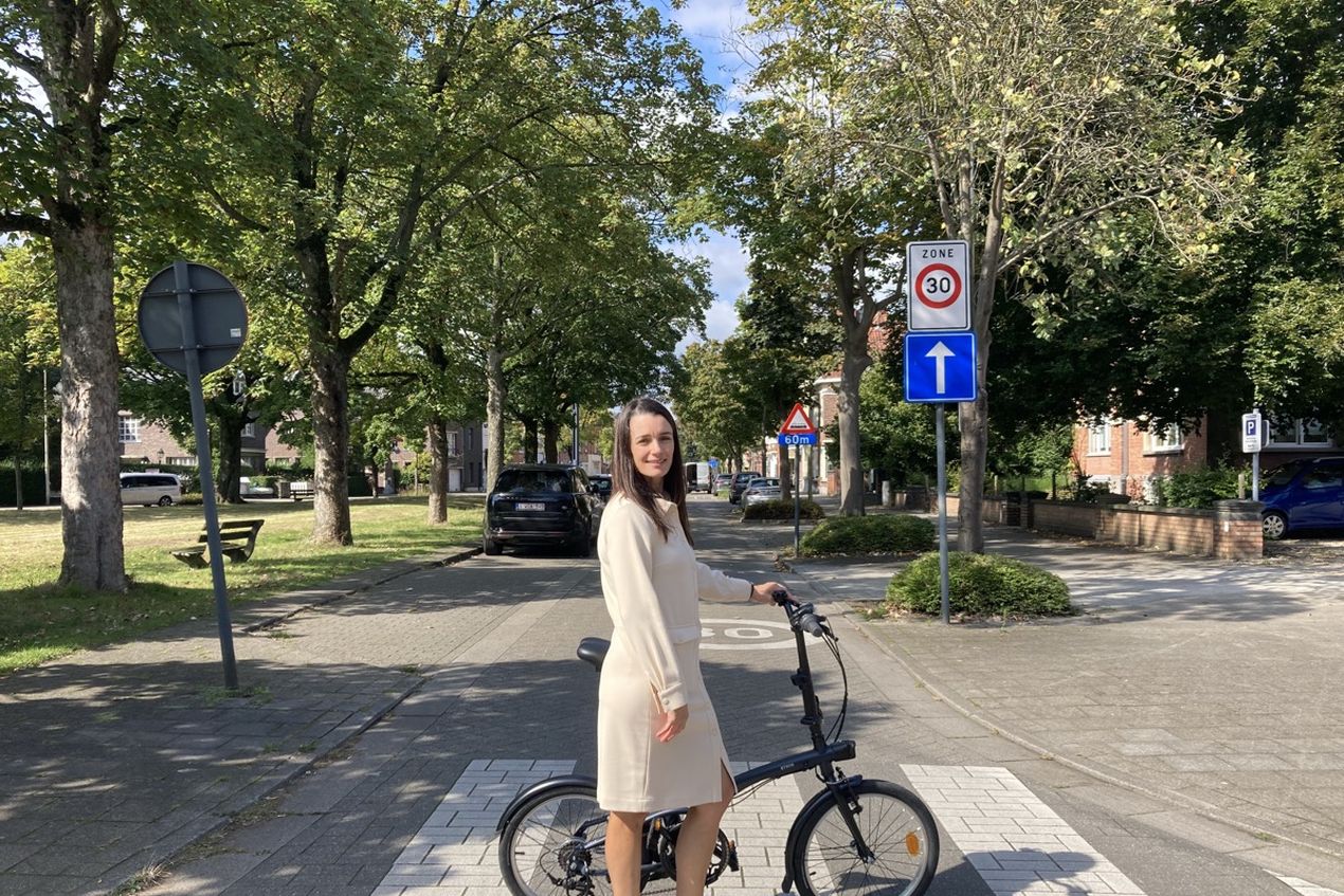 A woman in a white outfit stands on a pedestrian crossing with a folding bike, surrounded by trees and road signs in a suburban street.