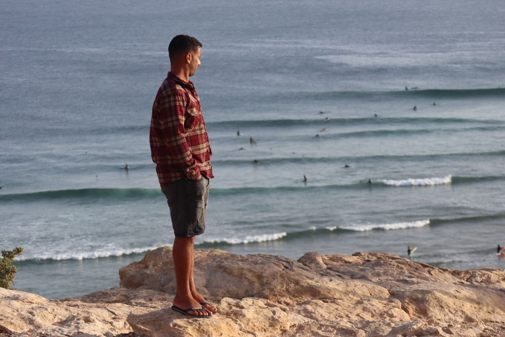 A person in a plaid shirt stands on a rocky cliff, overlooking the ocean with surfers catching waves below.