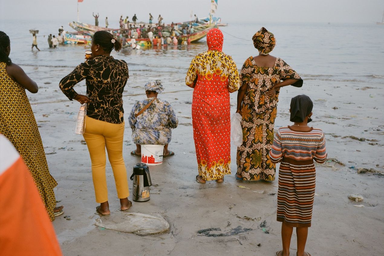 People in vibrant attire stand on a beach, observing a busy scene of colorful boats and activity in the water near the shore.