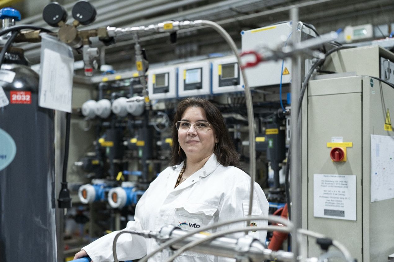 A woman in a lab coat stands in an industrial lab with complex machinery and instruments in the background.