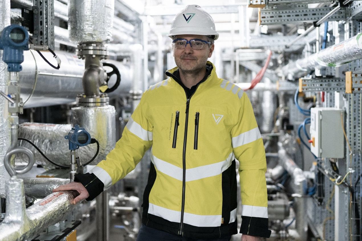 A person in a yellow safety jacket and hard hat stands in an industrial facility surrounded by metal pipes and equipment.