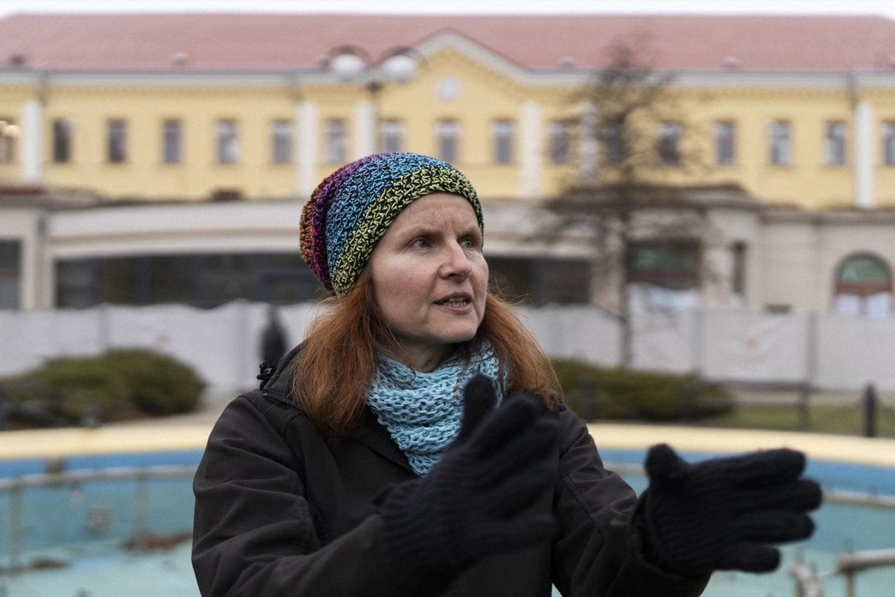 Woman in colorful knit hat and gloves gestures outdoors, with a yellow building and empty fountain in the background.