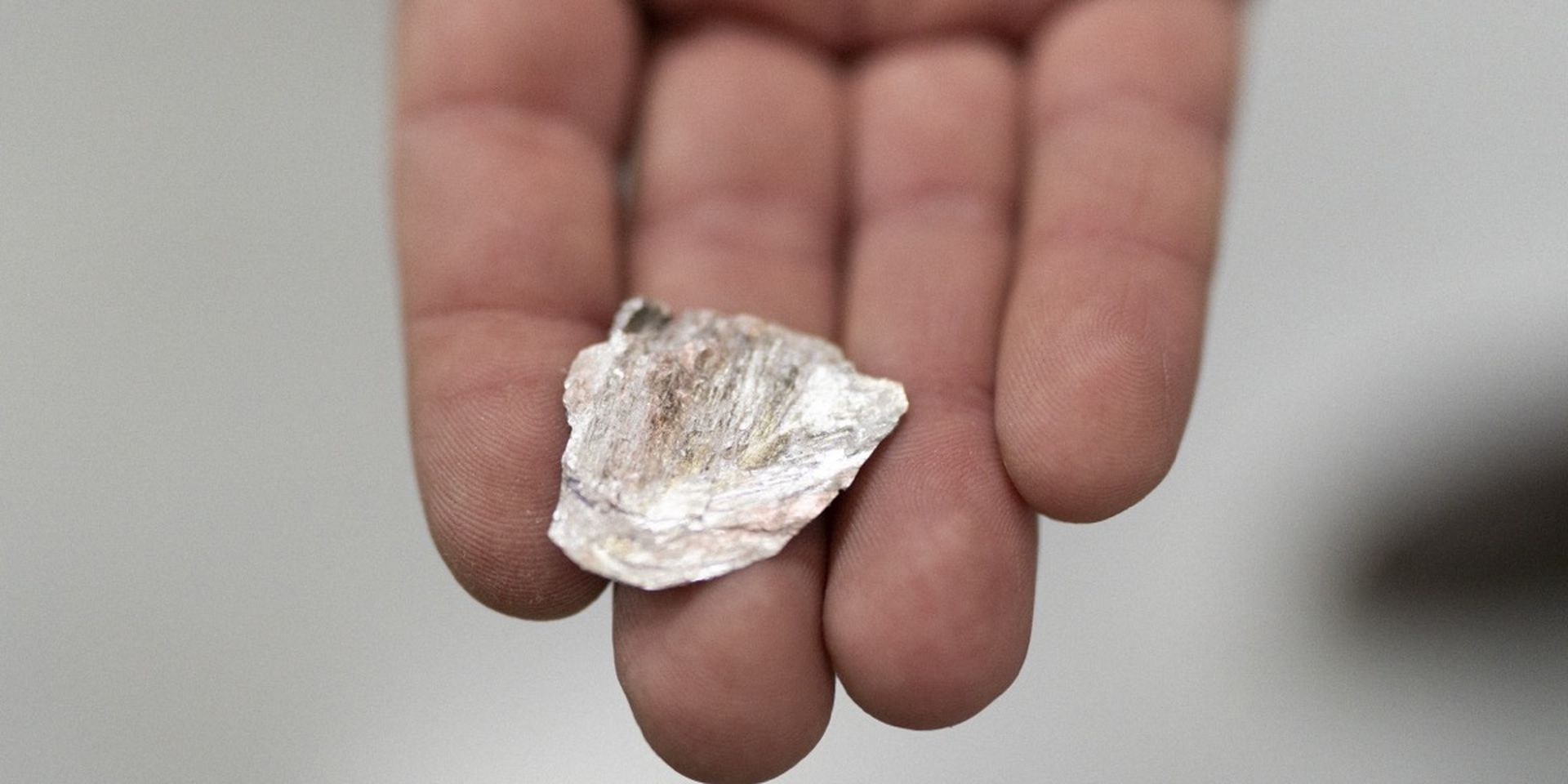 A hand holding a small, irregularly shaped shell with a rough texture and pinkish-white coloring against a blurred background.