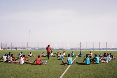 A group of soccer players stretch in a circle on a field, wearing blue and white jerseys, under a clear sky.