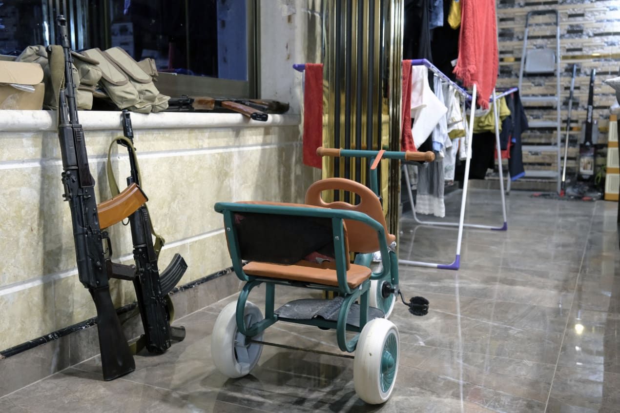 A children's tricycle sits in a room with rifles resting against a wall, alongside clothes drying on racks.