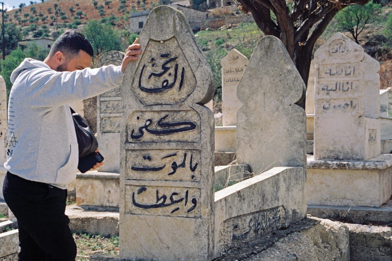 Man in a gray hoodie stands by an Arabic-inscribed gravestone in a cemetery with hills and trees in the background.
