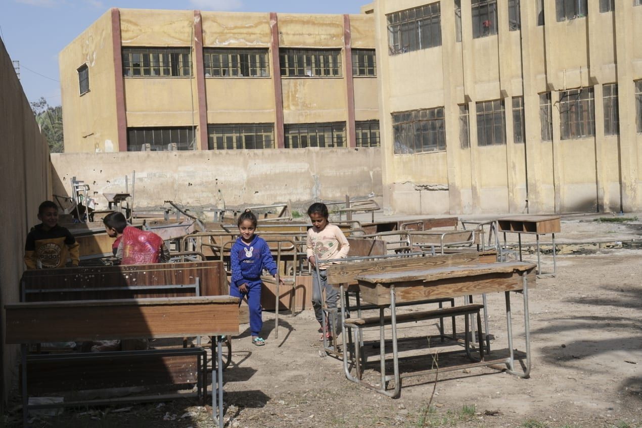 Children play among old desks in a schoolyard with a worn building in the background under a clear sky.