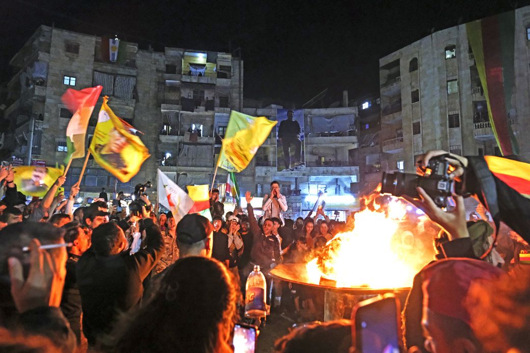 People gather at night, waving colorful flags around a large fire, with buildings in the background and someone taking photos.