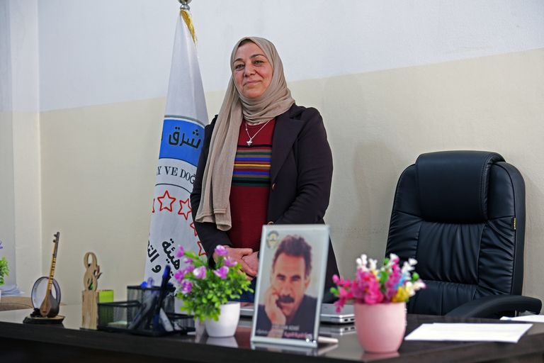 A woman in a hijab stands confidently in an office, with a flag and a desk adorned with flowers and a framed photo.
