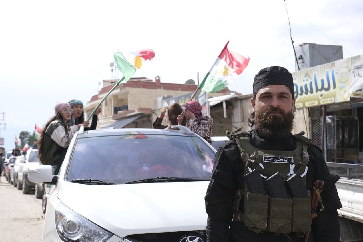 Bearded man in tactical gear stands beside a car as people wave Kurdish flags through the sunroof in an urban street scene.