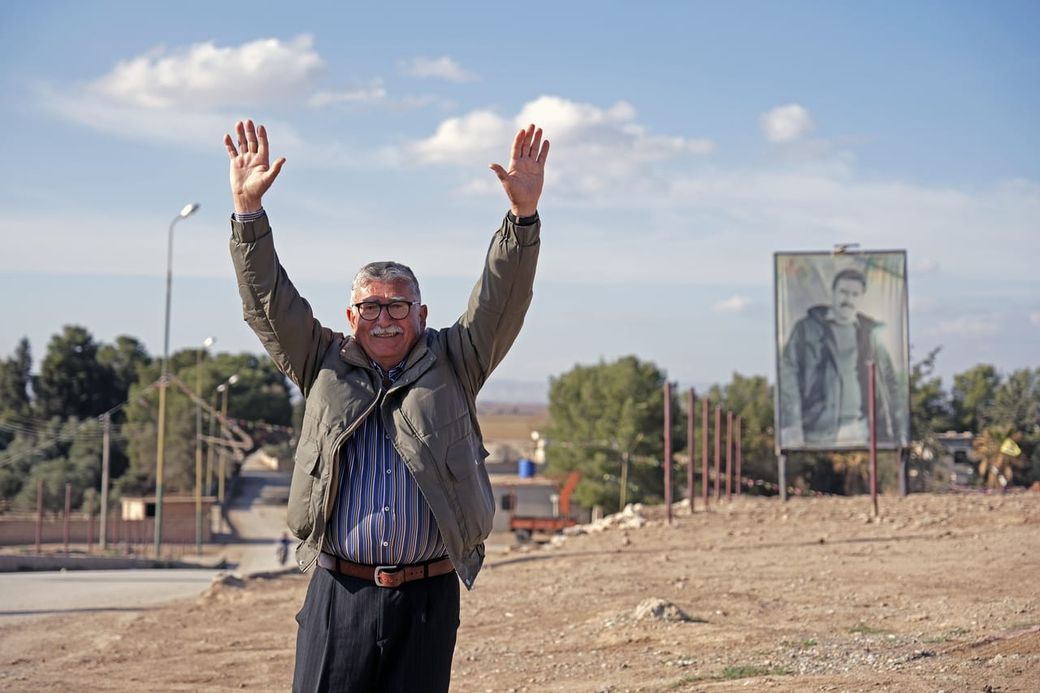 Smiling man with glasses raises arms joyfully outdoors; trees and a large portrait of a man in the background under a partly cloudy sky.