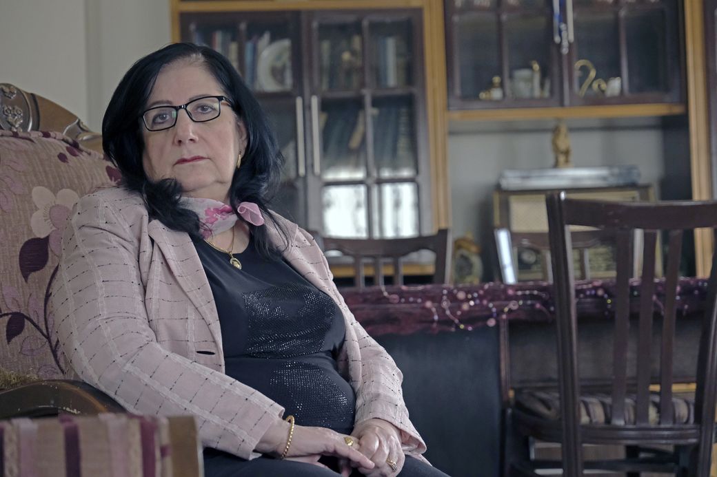 Woman with glasses and dark hair sitting on a floral chair, wearing a light pink jacket and scarf, in a room with a cabinet in the background.