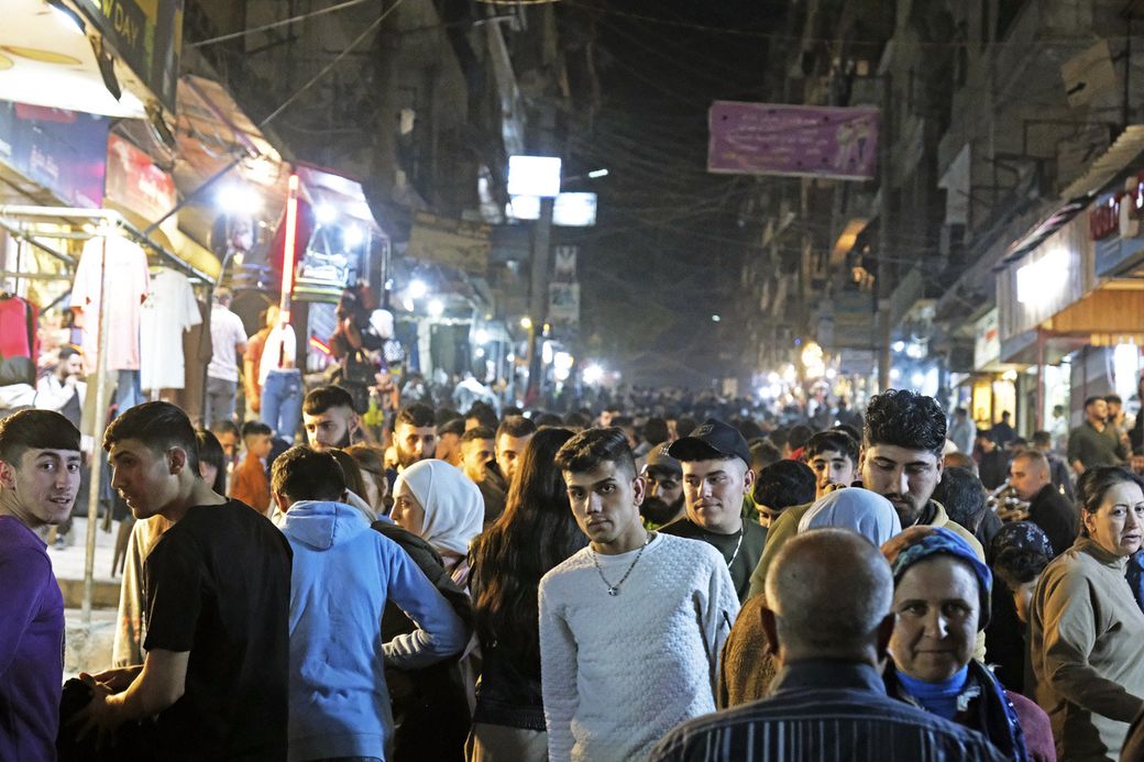A bustling market street at night, crowded with people walking and shopping under brightly lit vendor stalls.