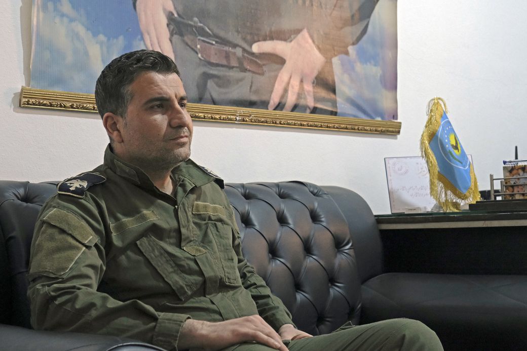A man in military uniform sits on a black leather sofa, with a large framed poster and a small blue and yellow flag in the background.